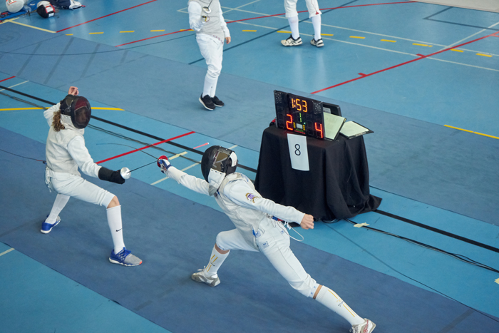 A fencing competition taking place with two fencer competing. One fencer is lunging forward with a strike while the other is defending